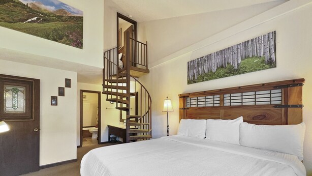 Ein Schlafzimmer mit einem großen Bett, einem hölzernen Kopfteil, einem Gemälde von Birken, einer Wendeltreppe und einer Tür mit einem Buntglasfenster.