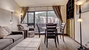 Ein gemütliches Wohnzimmer mit einer Couch, einem Couchtisch und einem Essbereich mit Stühlen und einem Tisch. Große Fenster bieten Blick auf Gebäude und Bäume.