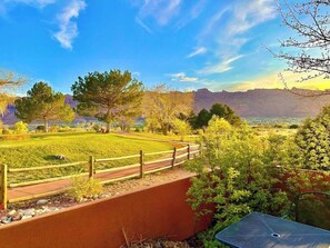 A fenced outdoor area with a paved path, trees, and a view of distant mountains. - Redesigned Red Rock condo with golf-course-view patio, fireplace, AC & pool (Moab)