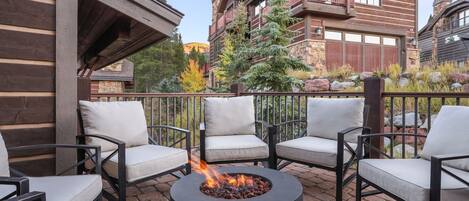 A patio area with a circular fire pit surrounded by four chairs, a wooden building with a balcony, and a stone building in the background.