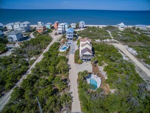 An aerial view of a coastal residential area with multiple houses, a central house with a swimming pool, and a beach in the background.