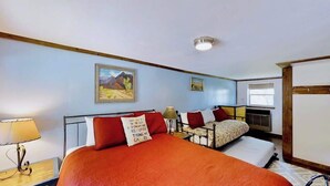 Habitación de hotel con una cama grande, una cama más pequeña, una mesita de noche con una lámpara y un cuadro en la pared.