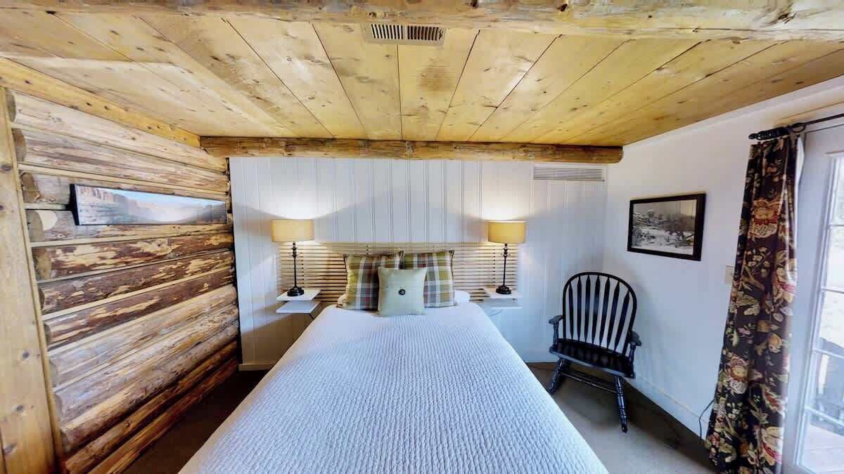 Um quarto com teto de madeira, uma cama com colcha branca, duas luminárias de cabeceira, uma cadeira e uma janela com cortinas.