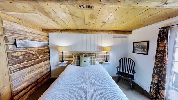 Um quarto com teto de madeira, uma cama com colcha branca, duas luminárias de cabeceira, uma cadeira e uma janela com cortinas.
