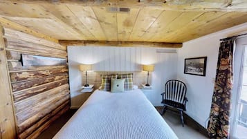 Um quarto com teto de madeira, uma cama com colcha branca, duas luminárias de cabeceira, uma cadeira e uma janela com cortinas.