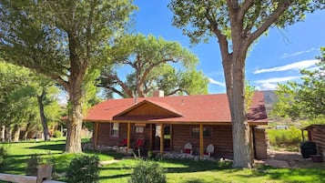 Uma cabana rústica de madeira com telhado vermelho, rodeada por vegetação exuberante e árvores grandes.