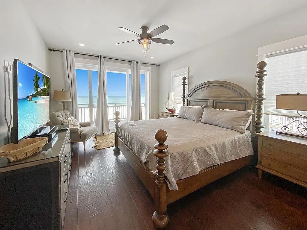 A bedroom with a large bed, a ceiling fan, a TV, and a view of the ocean through the window.
