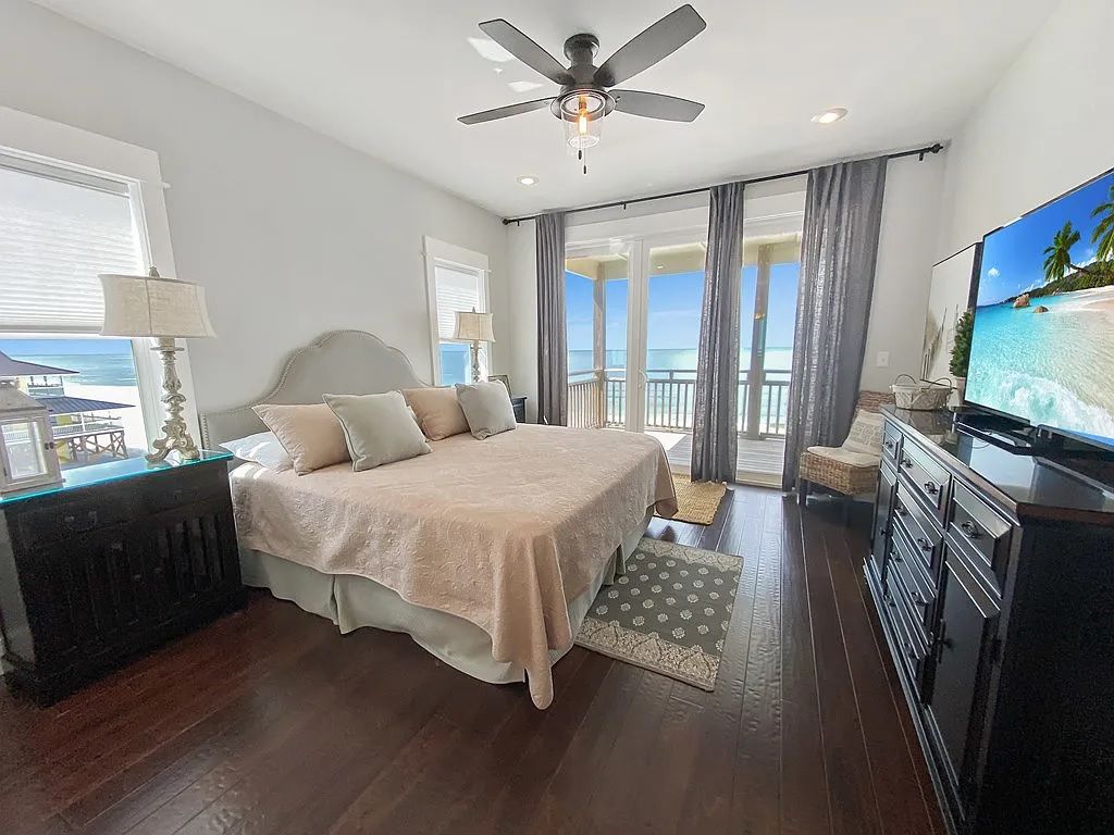 A bedroom with a bed, a television, a ceiling fan, and a view of the ocean through large windows.
