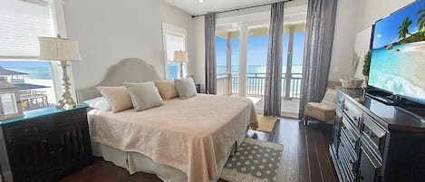 A bedroom with a bed, a television, a ceiling fan, and a view of the ocean through large windows.