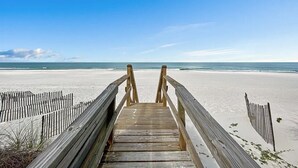 Ein hölzerner Steg führt zu einem Sandstrand unter blauem Himmel.