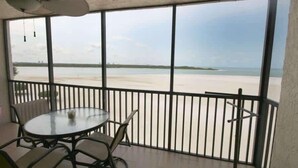 A balcony with a glass table and chairs, a fan, and a view of a beach. - Carlos Pointe #531 Cozy Beachfront Condo (Fort Myers Beach)