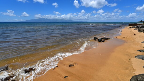 Ein Strand mit klarem, blauem Wasser, sandigem Ufer und vereinzelten Felsen.