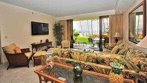 A spacious living room with a large sofa, a coffee table, and a television mounted on the wall. There is a view of a tropical garden and the ocean through large windows. - Time to Spoil Yourself!  Deluxe Ocean Front and Steps to N. Kaanapali Beach ~ (Lahaina)