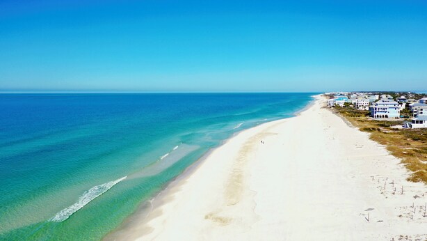 Une plage c ôtière aux eaux turquoise et limpides, bordée d’un rivage sablonneux. Des bâtiments sont construits le long de la plage, ce qui laisse supposer qu’il s’agit d’une station balnéaire ou d’une zone de villégiature.