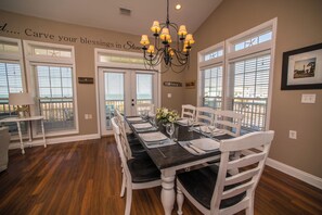 A dining area with a long table set for four, white chairs, a chandelier, and a wall with a decorative phrase.