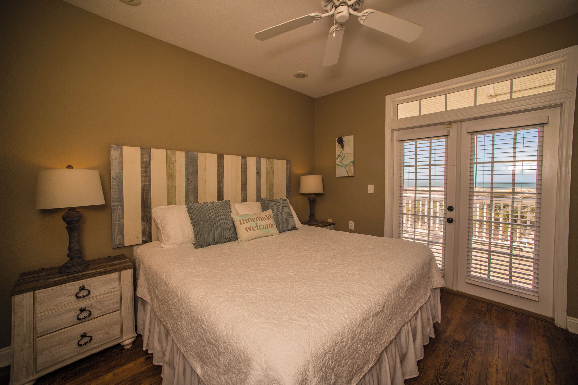 A bedroom with a large bed, two bedside lamps, a ceiling fan, a wooden nightstand, and a view through the door.