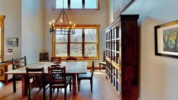 A dining area with a large wooden table and chairs, a chandelier, a window with a view of trees, and a bookshelf.