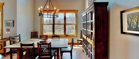 A dining area with a large wooden table and chairs, a chandelier, a window with a view of trees, and a bookshelf.