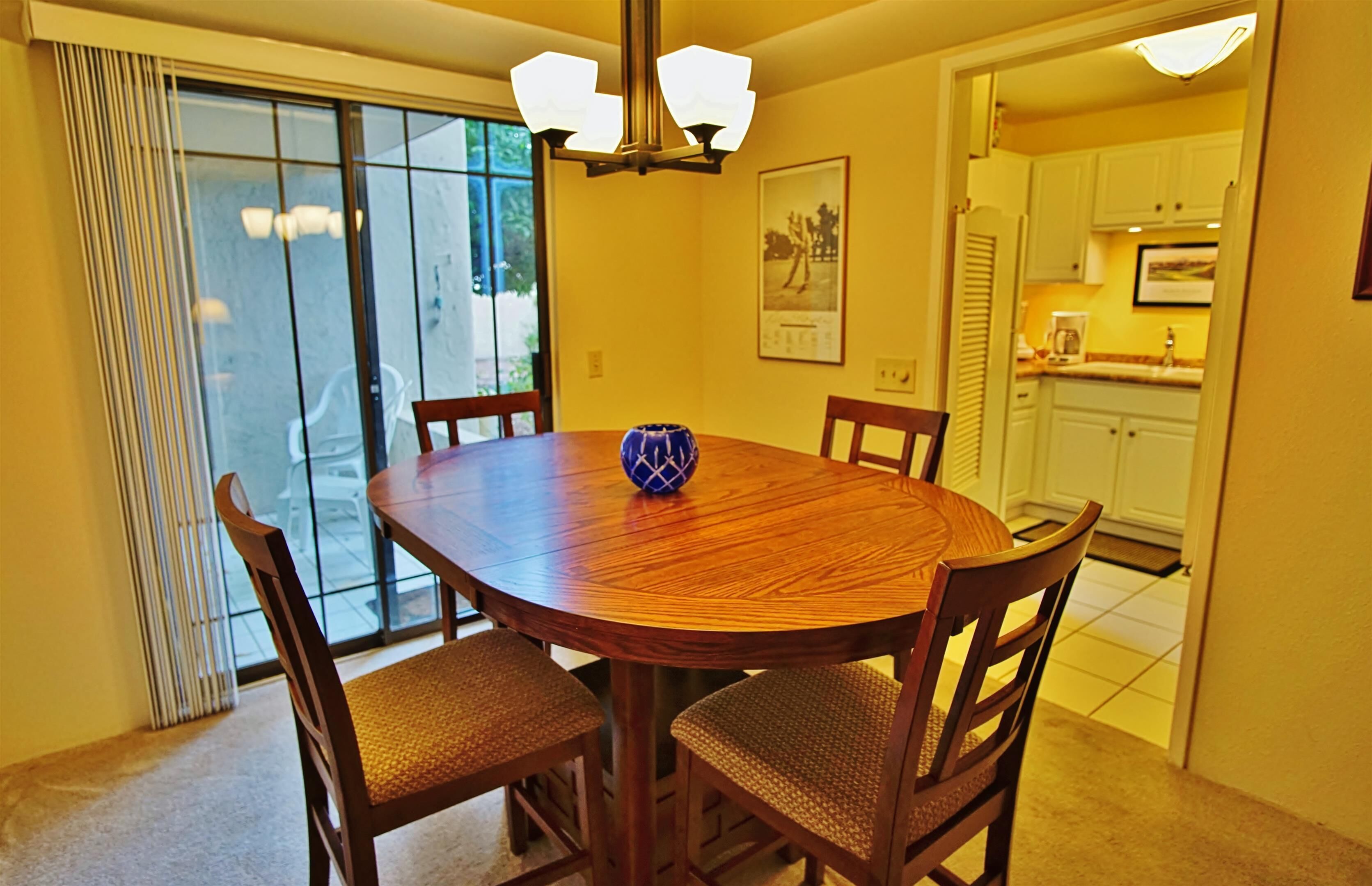 A dining area with a round wooden table, four chairs, and a blue vase. There is a sliding glass door leading to an outdoor area.