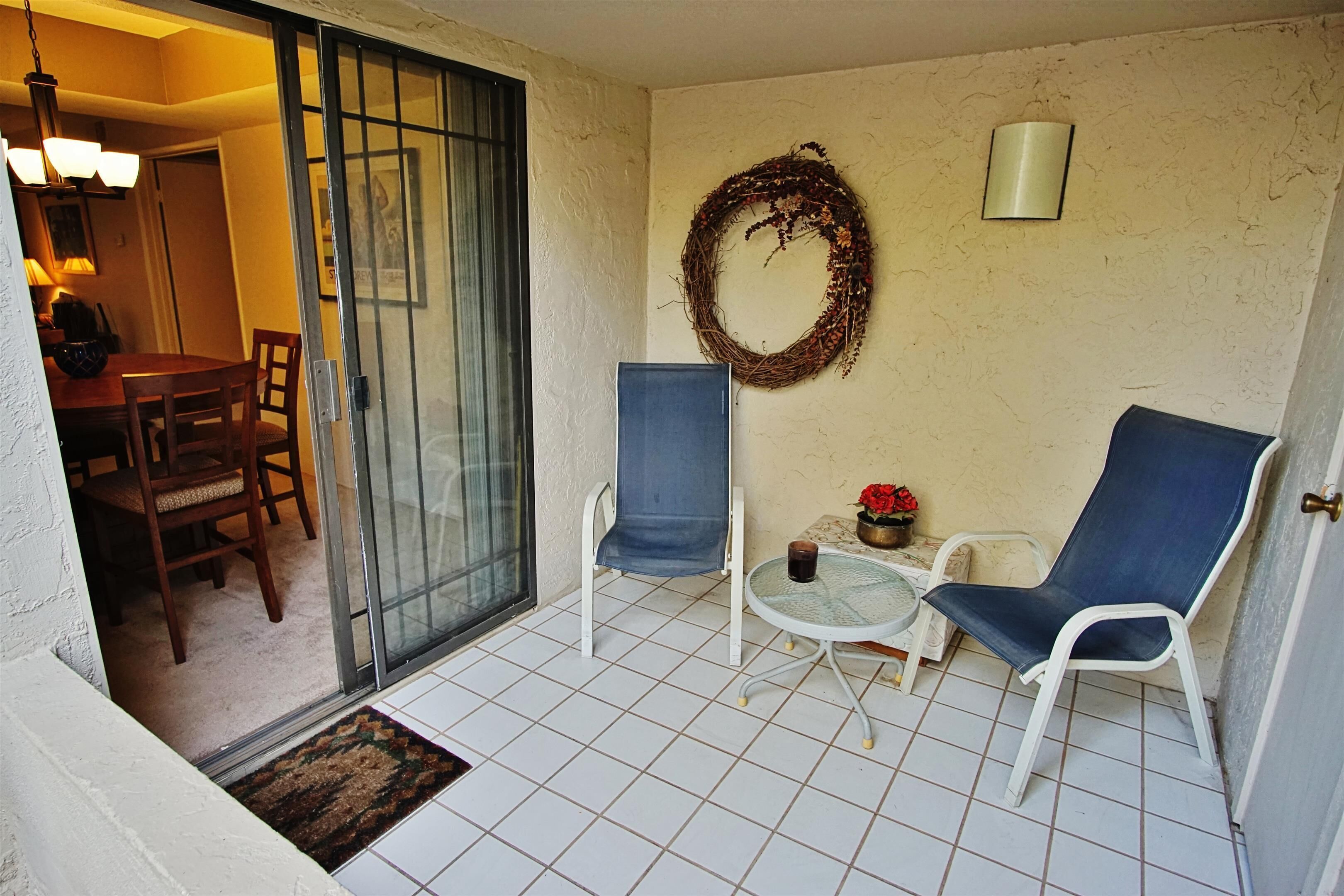 A balcony with two chairs, a small table, a decorative wreath, and a sliding glass door leading to a dining area.