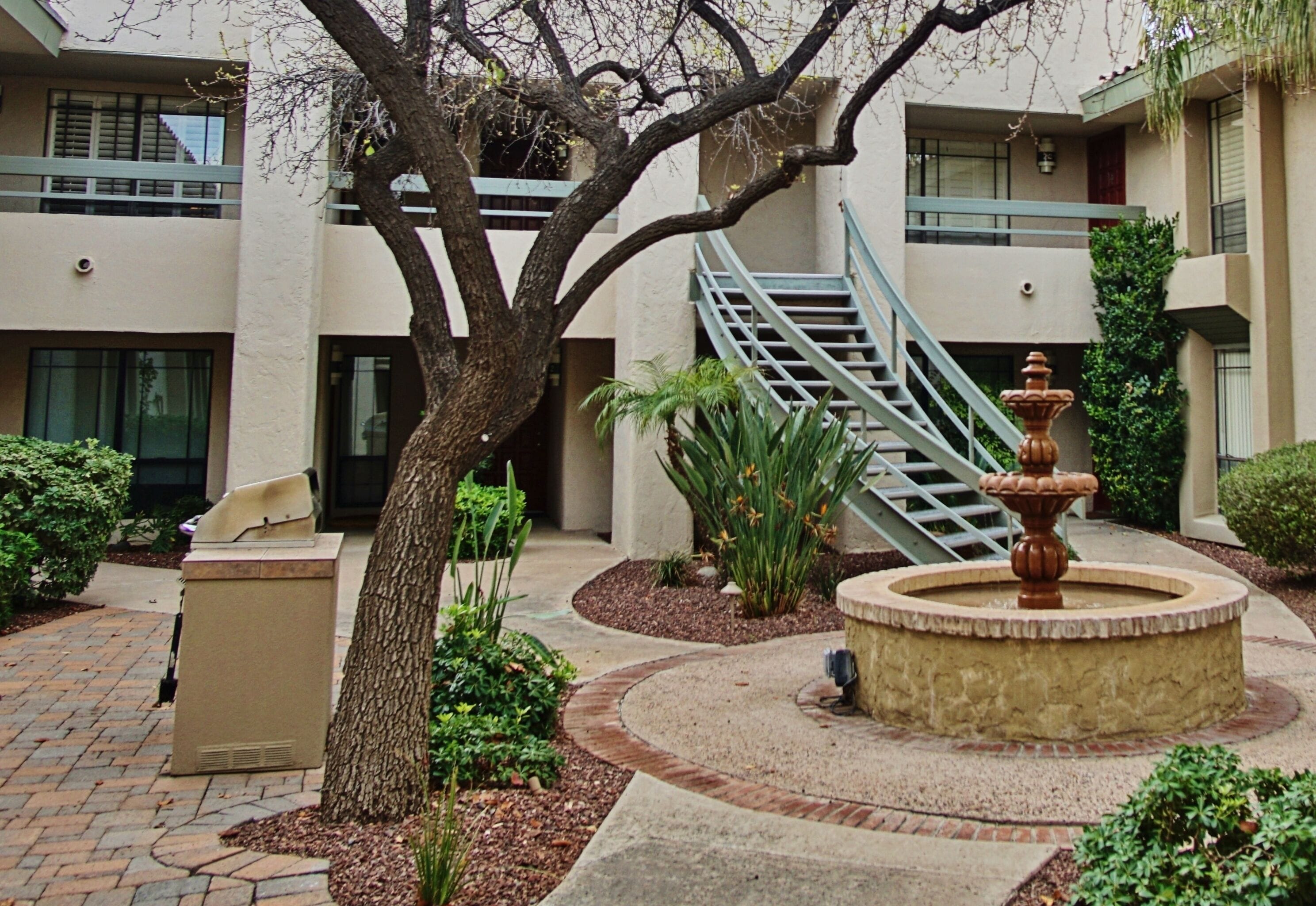 Une cour intérieure avec une fontaine, un arbre et un escalier menant à un niveau supérieur.