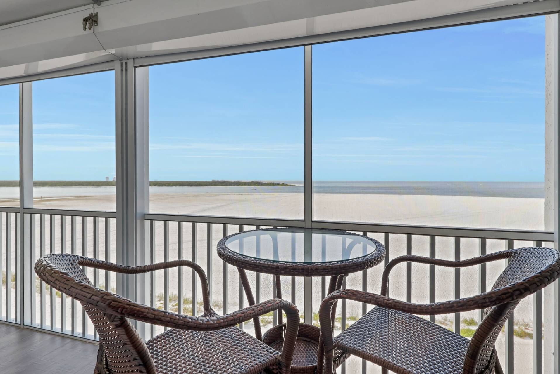A balcony with wicker chairs and a glass table, overlooking a beach with a clear sky.