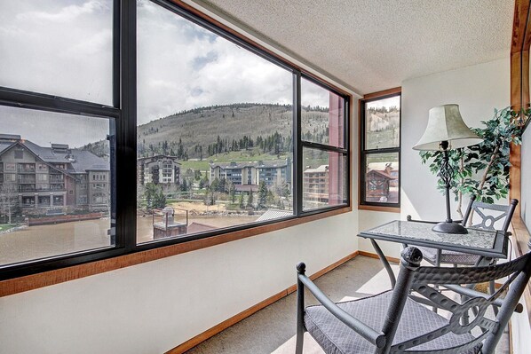 A room with large windows offering a view of a mountainous area and buildings. There is a table with a lamp and a chair.