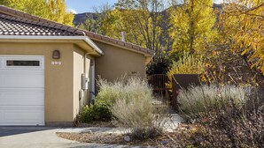 A beige house with a tiled roof, a white garage door, and a small garden with shrubs and trees. - Downtown Location | Indoor Pool | Fenced Backyard (Moab)