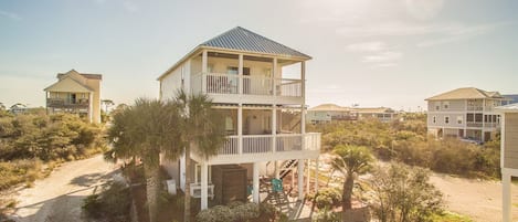 Una casa frente al mar de varios pisos, con balcón, rodeada de vegetación y otras casas parecidas.