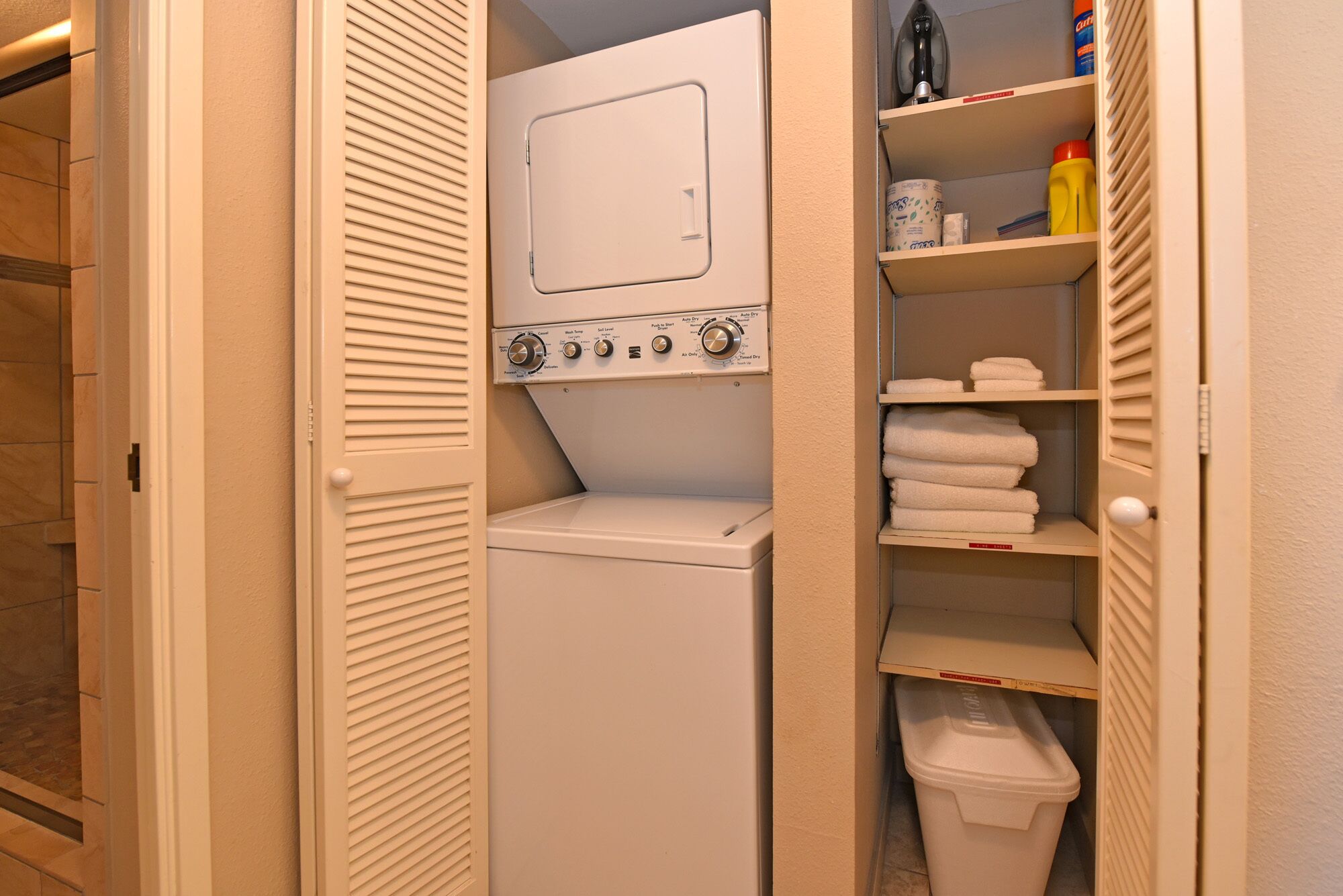 Una sala de lavandería con lavadora, estantes con toallas y un basurero.