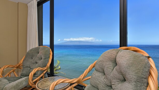Un balcone con sedie in vimini e un tavolino rotondo, con vista su un mare azzurro e un'isola in lontananza.