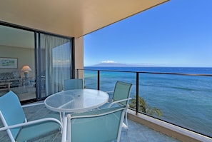 Un balcón con una mesa y sillas de vidrio, con vista a un mar azul y montañas lejanas.
