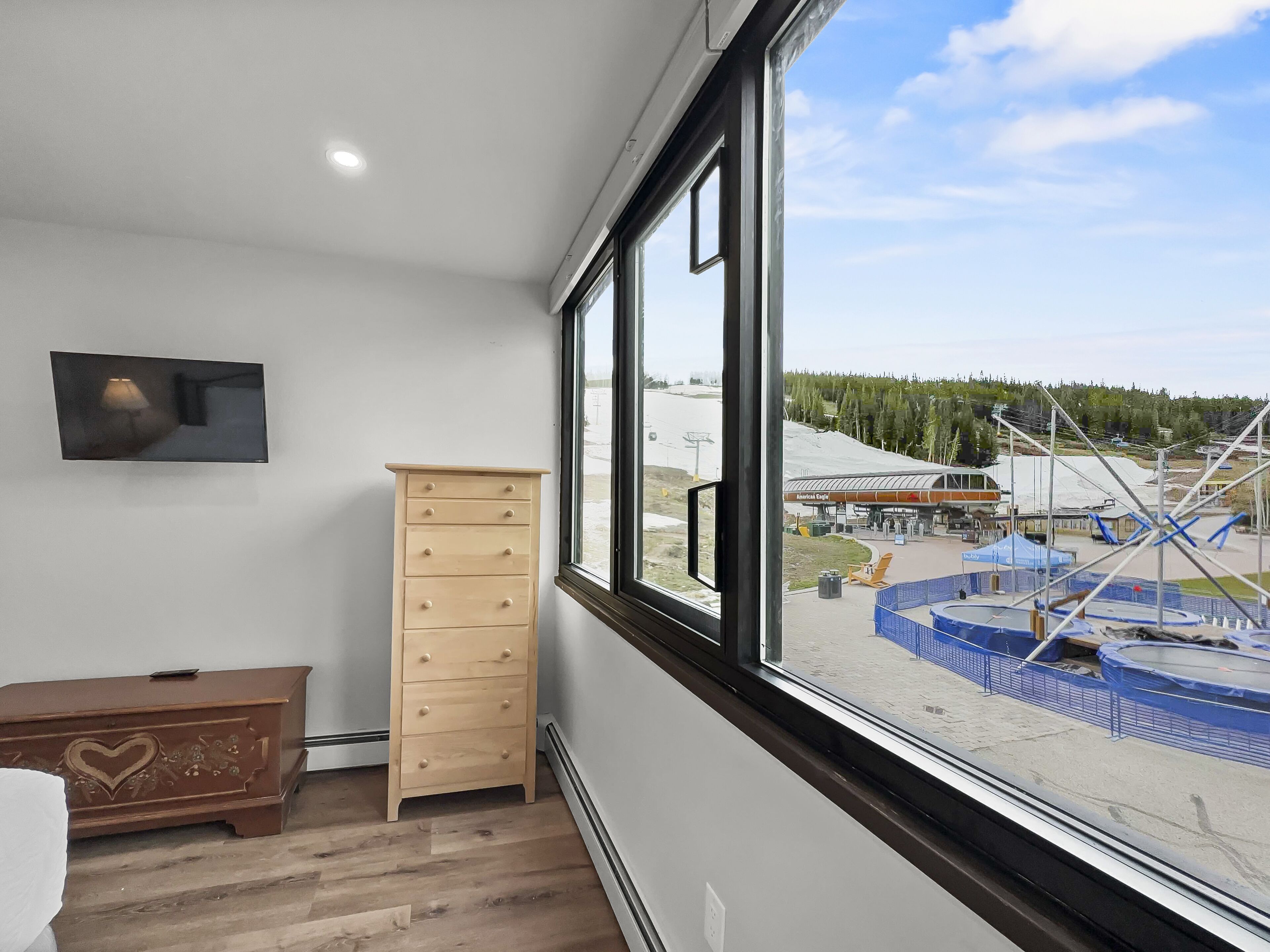 A room with a wooden dresser, a television mounted on the wall, and a large window offering a view of an outdoor area with a gas station and construction site.