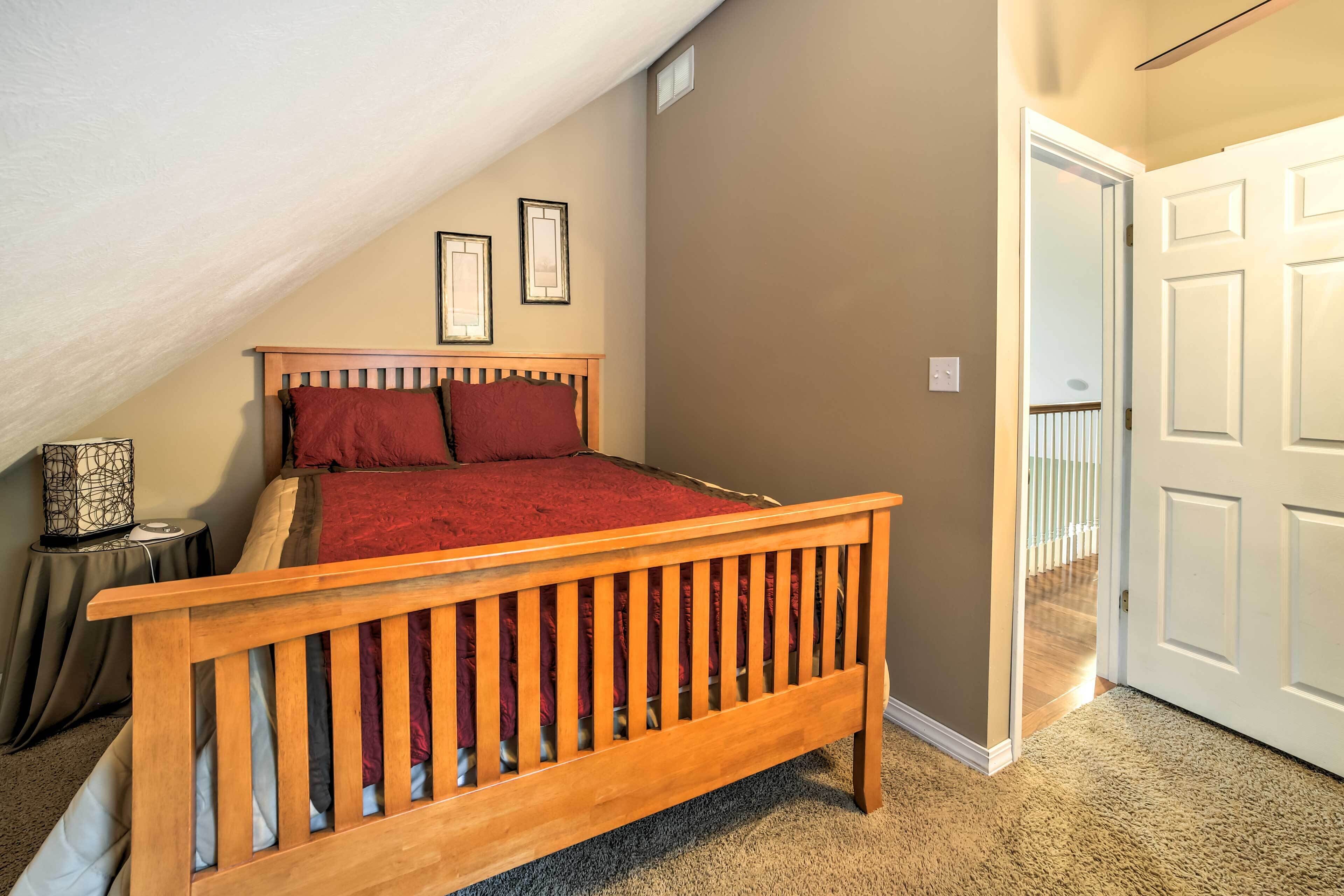 Une chambre avec un plafond en pente, un lit en bois recouvert d’une couverture rouge, une table de chevet avec une lampe et une porte donnant sur un escalier.