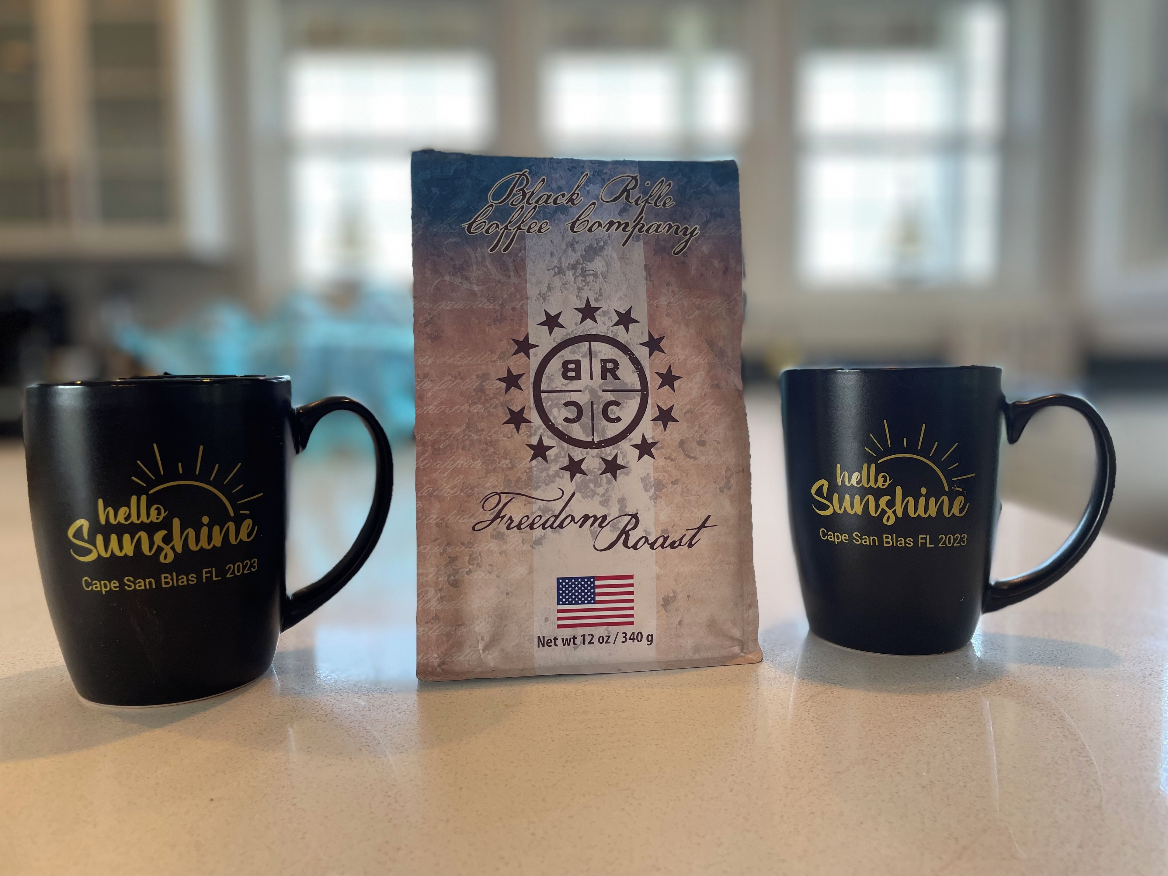 Two black coffee mugs with 'hello Sunshine Cape San Blas FL 2023' text, a coffee bag with 'Black Rifle Coffee Company Freedom Roast' and 'BRIC' logo, and an American flag.