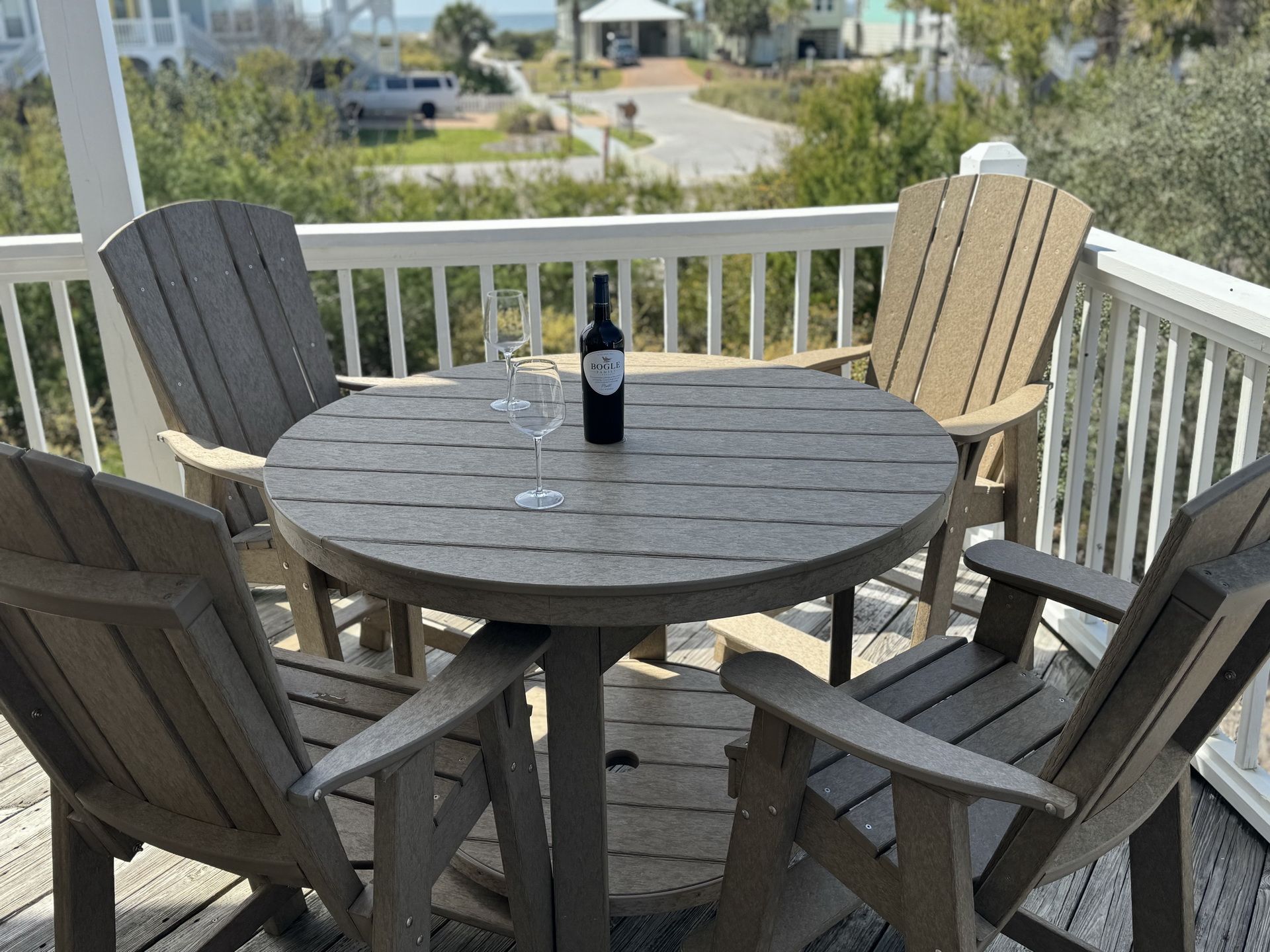 Et rundt træbord med fire stole på en terrasse, en vinflaske og to glas.