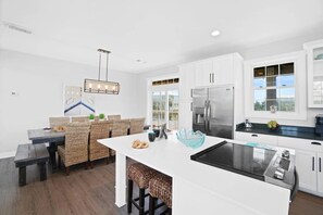 A modern kitchen with white cabinetry, a stainless steel refrigerator, a built-in stove, and a dining area with wicker chairs and a bench.