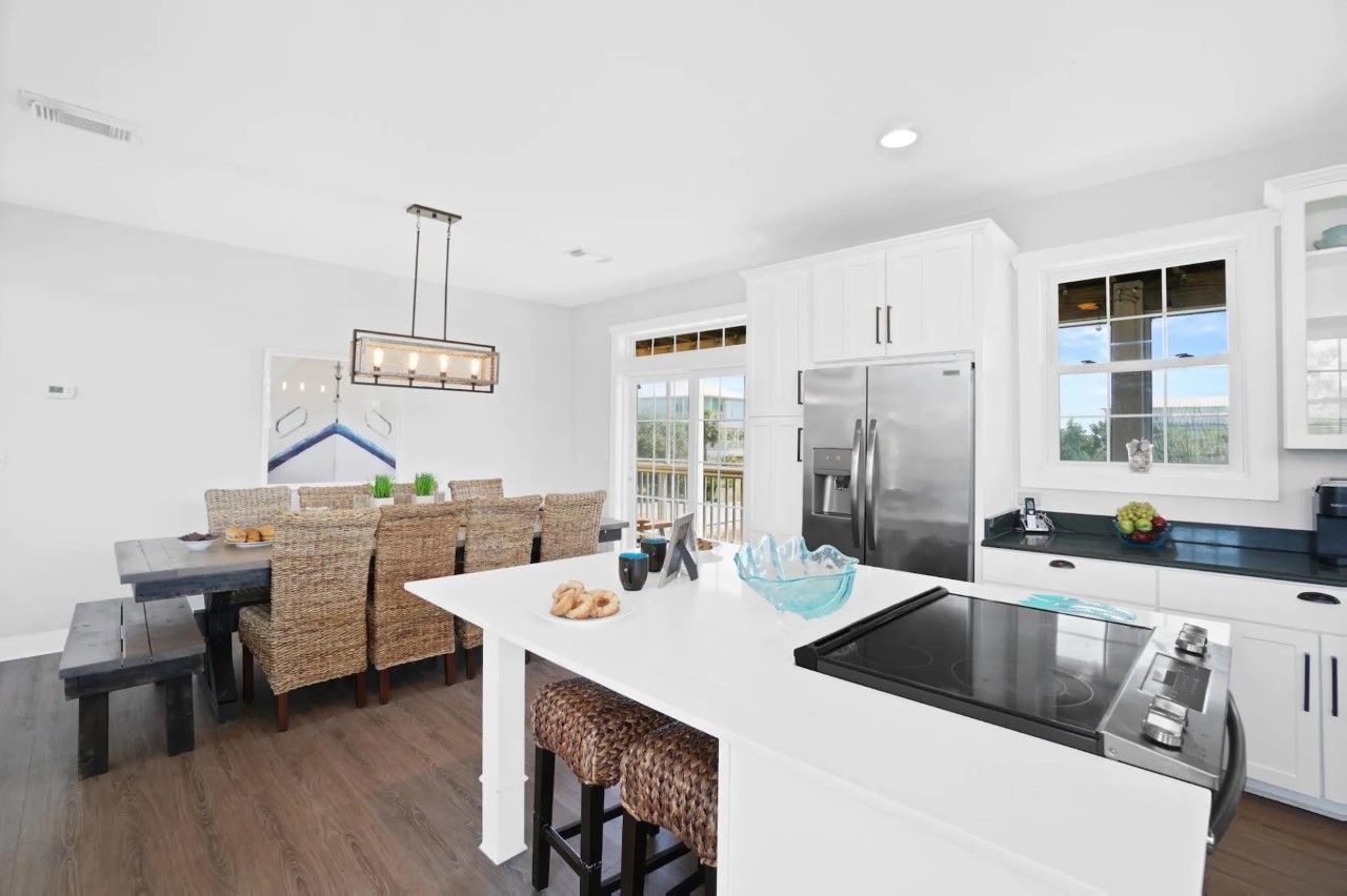 A modern kitchen with white cabinetry, a stainless steel refrigerator, a built-in stove, and a dining area with wicker chairs and a bench.