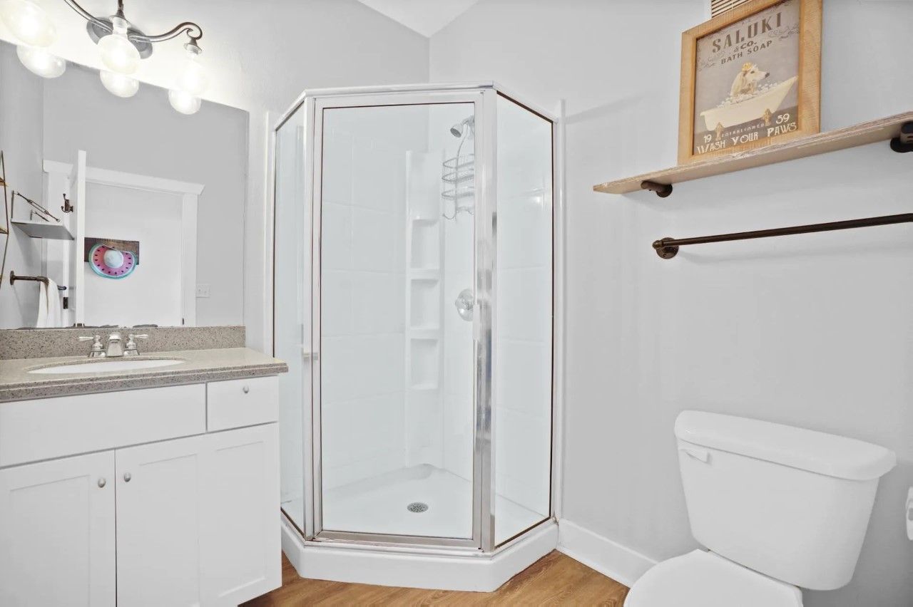 Un baño en esquina con ducha envuelta en vidrio, inodoro blanco, lavabo con encimera y un anuncio enmarcado en la pared.