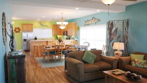 Amplia sala de estar con sofá, mesa de centro y una mesa de comedor lista para cuatro personas. Se ve una cocina con armarios de madera y un refrigerador al fondo.