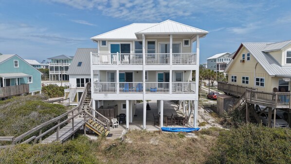 A multi-story beachfront building with balconies, a covered lower level, and a wooden deck leading to the entrance.