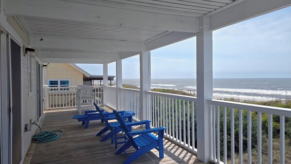 A covered porch with a view of the ocean, featuring a wooden deck and blue chairs.