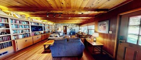 L’intérieur chaleureux d’un chalet avec un plafond en bois, une cheminée, un téléviseur et une bibliothèque.