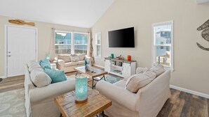 A spacious living room with a high ceiling, a flat-screen TV mounted on the wall, and a large window providing a view of the cityscape. - 2 minute walk to the beach, stores and restaurants (Cape San Blas)