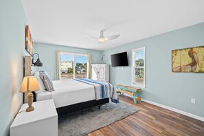 A bedroom with a bed, a nightstand, a lamp, a ceiling fan, a TV, and a window with a view of buildings. - 2 minute walk to the beach, stores and restaurants (Cape San Blas)