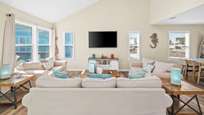 A spacious living room with a large sofa, a coffee table, and a TV mounted on the wall. There are windows with curtains, a bookshelf, and a dining area with a table and chairs. - 2 minute walk to the beach, stores and restaurants (Cape San Blas)