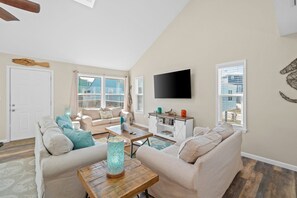 A spacious living room with a high ceiling, a flat-screen TV mounted on the wall, and a large window providing a view of the cityscape.