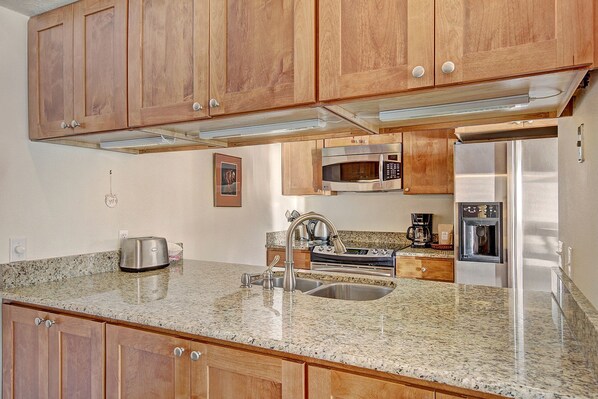 Una cocina con armarios de madera, encimera de granito y electrodomésticos de acero inoxidable, incluyendo un refrigerador, microondas y horno, además de una cafetera.