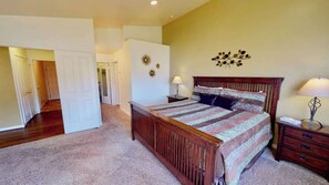 A bedroom with a wooden bed, two bedside tables, a nightstand, a wall-mounted lamp, and a door leading to another room. - Desert Hideaway - C2, Enjoy The Views (Moab)