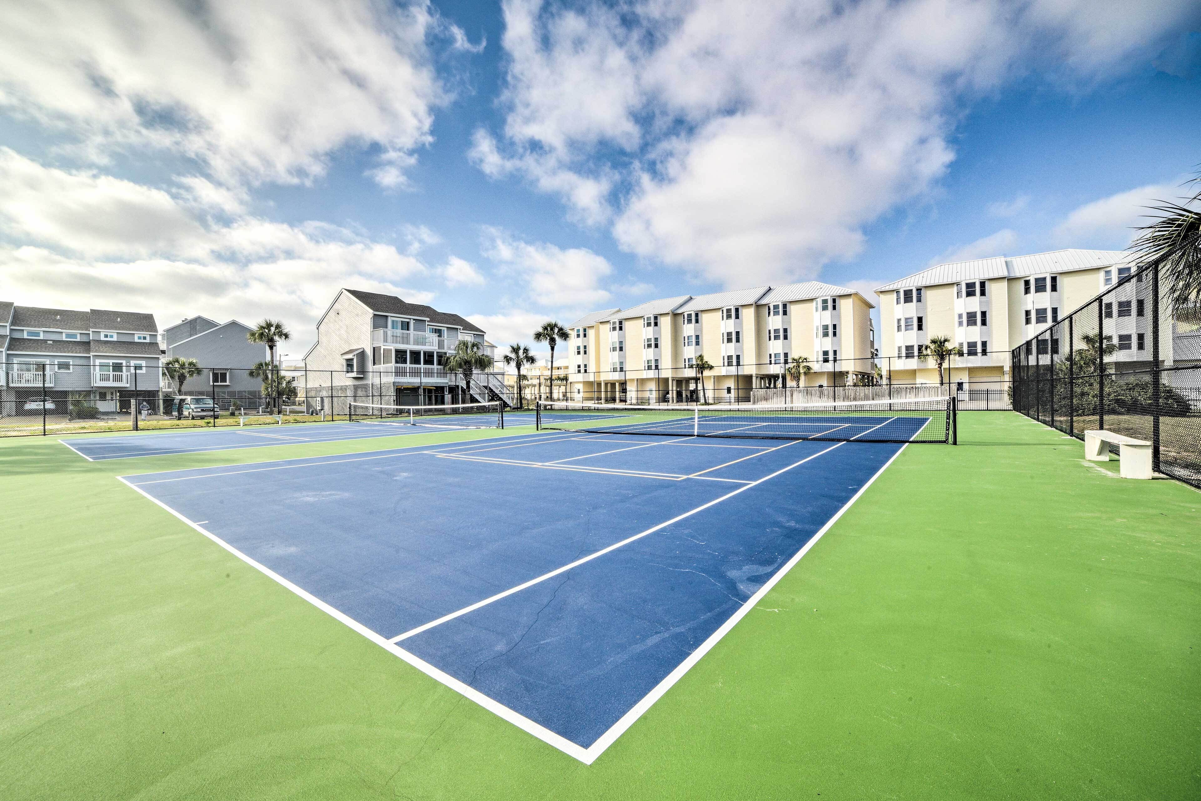 A tennis court with a net, surrounded by a fence and palm trees, with buildings in the background.